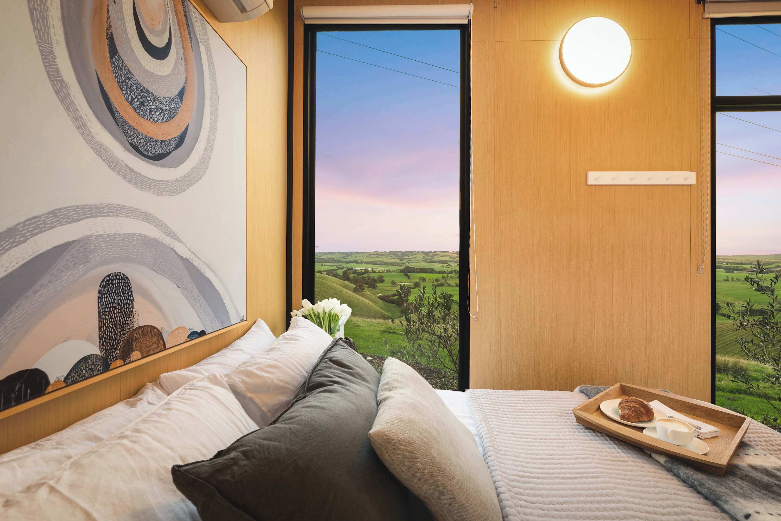 A bedroom with a bed, pillows, a tray with a croissant, and a view of a green landscape through a window.