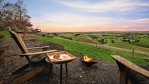 A rustic outdoor seating area with a fire pit, a tray with a croissant and cup, and a scenic view of rolling hills. - Belrose Estate by Tiny Away (Dumbalk)