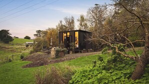 A small, dark-colored cabin with a visible entrance and a small porch area, surrounded by greenery and trees. - Belrose Estate by Tiny Away (Dumbalk)