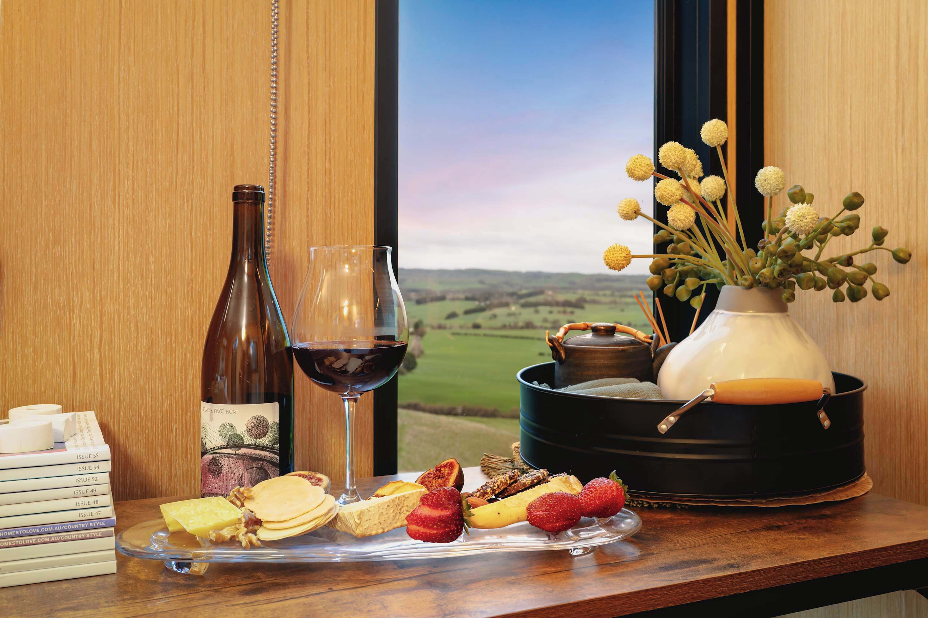 A table with a wine bottle, glass, cheese, fruits, and a vase with flowers.