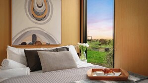 A hotel room with a large bed, a painting above it, a window with a view of greenery, and a tray with breakfast items. - Belrose Estate by Tiny Away (Dumbalk)