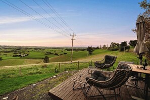 Una terraza de madera con sillas de mimbre y una mesa, con vistas a un paisaje verde con campos y árboles.