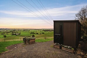 A small, dark-colored structure with a door, surrounded by a gravel path and wooden planters with greenery. - Belrose Estate by Tiny Away (Dumbalk)