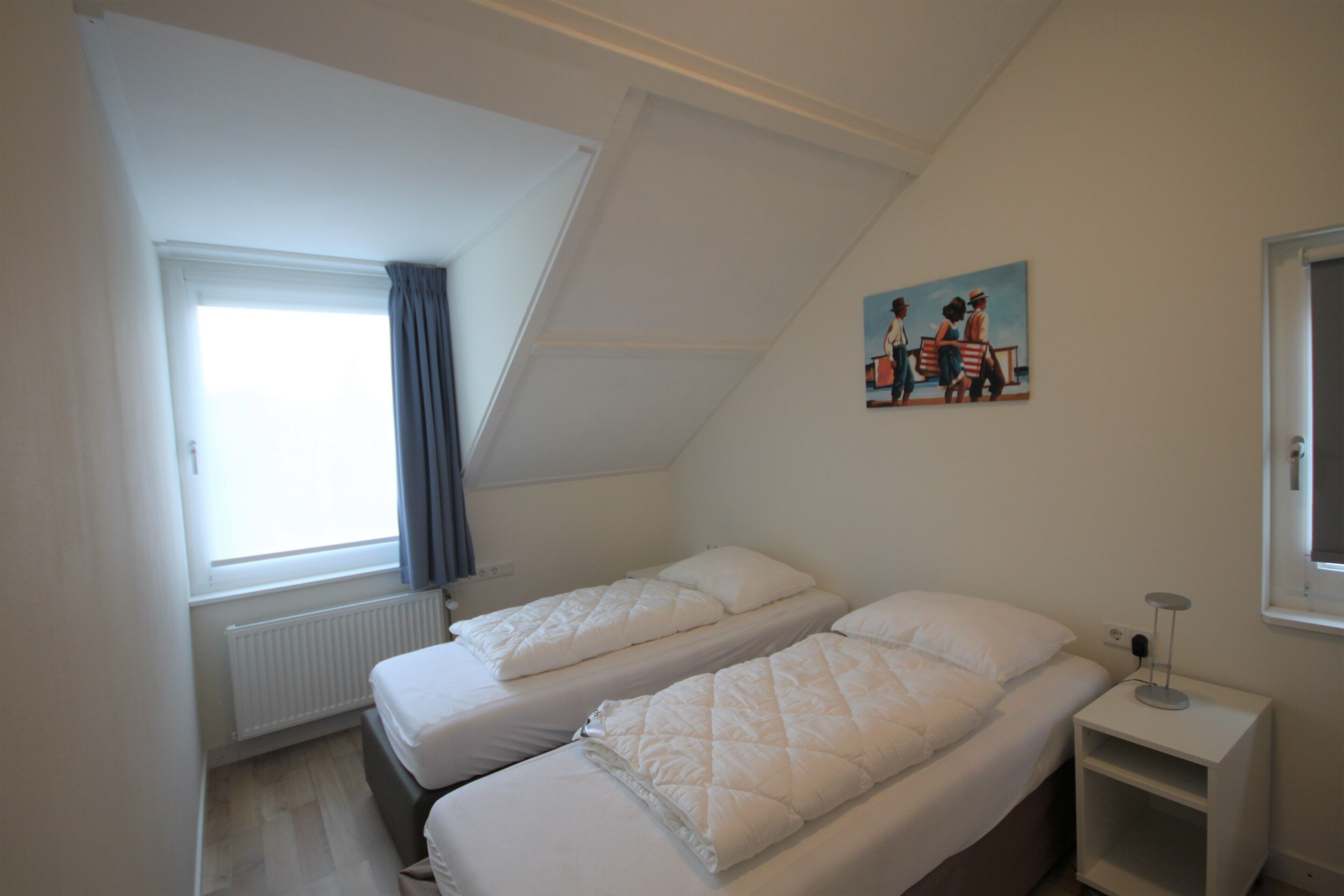 Una camera da letto con due letti, una finestra con tende, un termosifone, un quadro al muro e un comodino con una lampada.