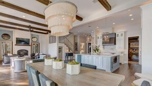 A spacious living area with a wooden dining table, comfortable seating, a chandelier, and a kitchen with white cabinets and a blue island.