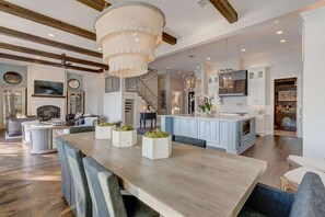 A spacious living area with a wooden dining table, comfortable seating, a chandelier, and a kitchen with white cabinets and a blue island.