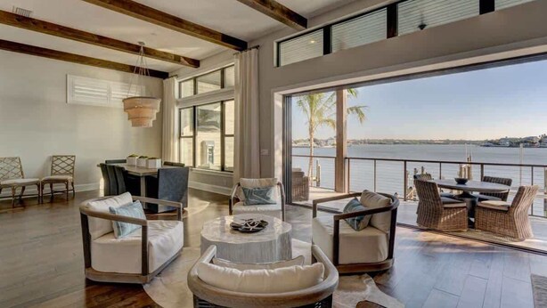 A spacious living area with a large window overlooking a body of water, furnished with wicker chairs and a round table.