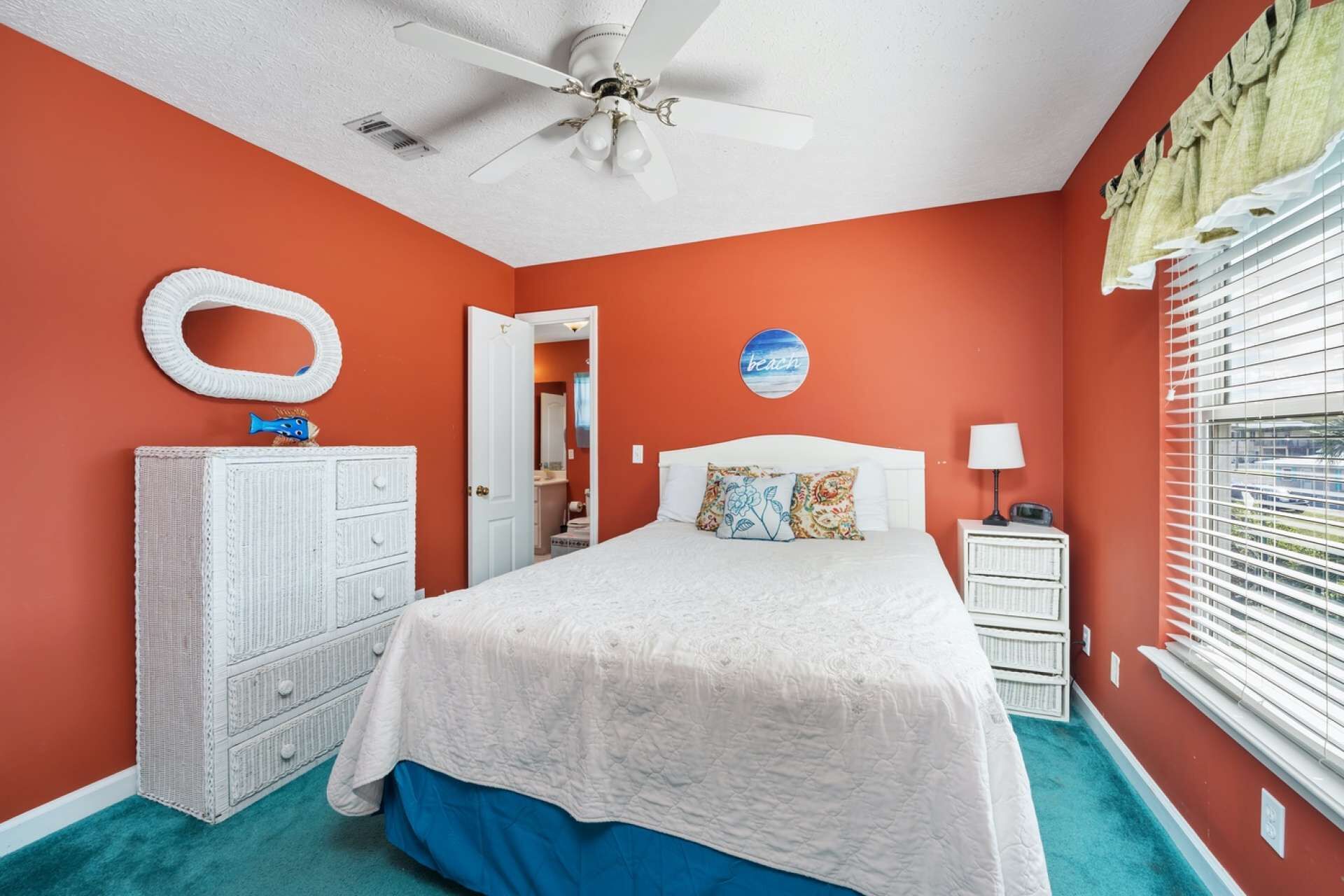 A bedroom with a white bed, a ceiling fan, a white dresser, a nightstand with a lamp, and a window with blinds.