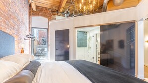 A modern bedroom with a brick wall, a large bed with white and gray bedding, a hanging light fixture, and a sliding glass door leading to a bathroom.