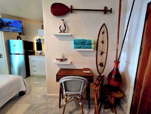 A hotel room with a bed, a desk with a chair, a television, a refrigerator, decorative items on the wall, and a window with curtains. - Oceanfront studio see whales and dolphins (Pāhoa)