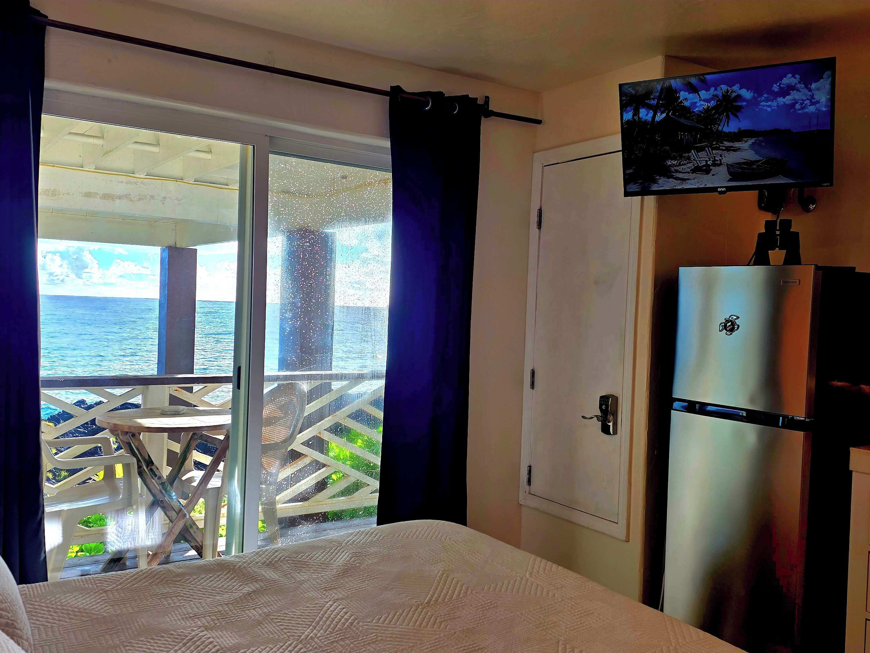A bedroom with a bed, a television mounted on the wall, and a sliding door leading to a balcony with a view of the sea.