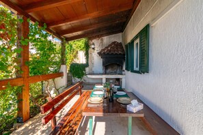 An outdoor dining area with a wooden table set for two, a bench, and a stone fireplace.