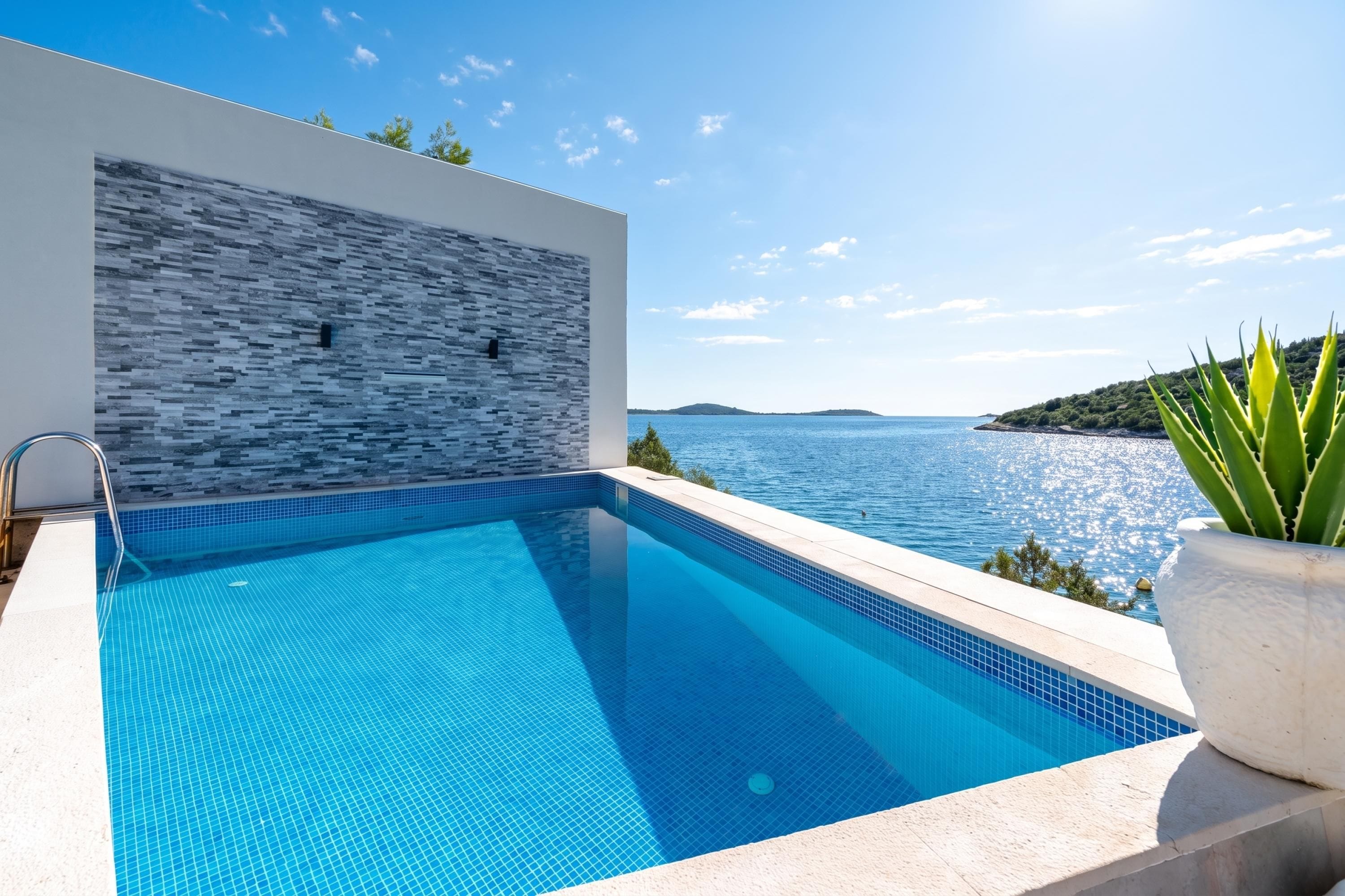 A swimming pool with a tiled edge, a white wall with a textured pattern, and a view of the sea and a distant island.