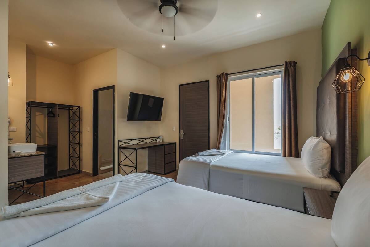 A hotel room with two beds, a desk, a TV, and a window with curtains.