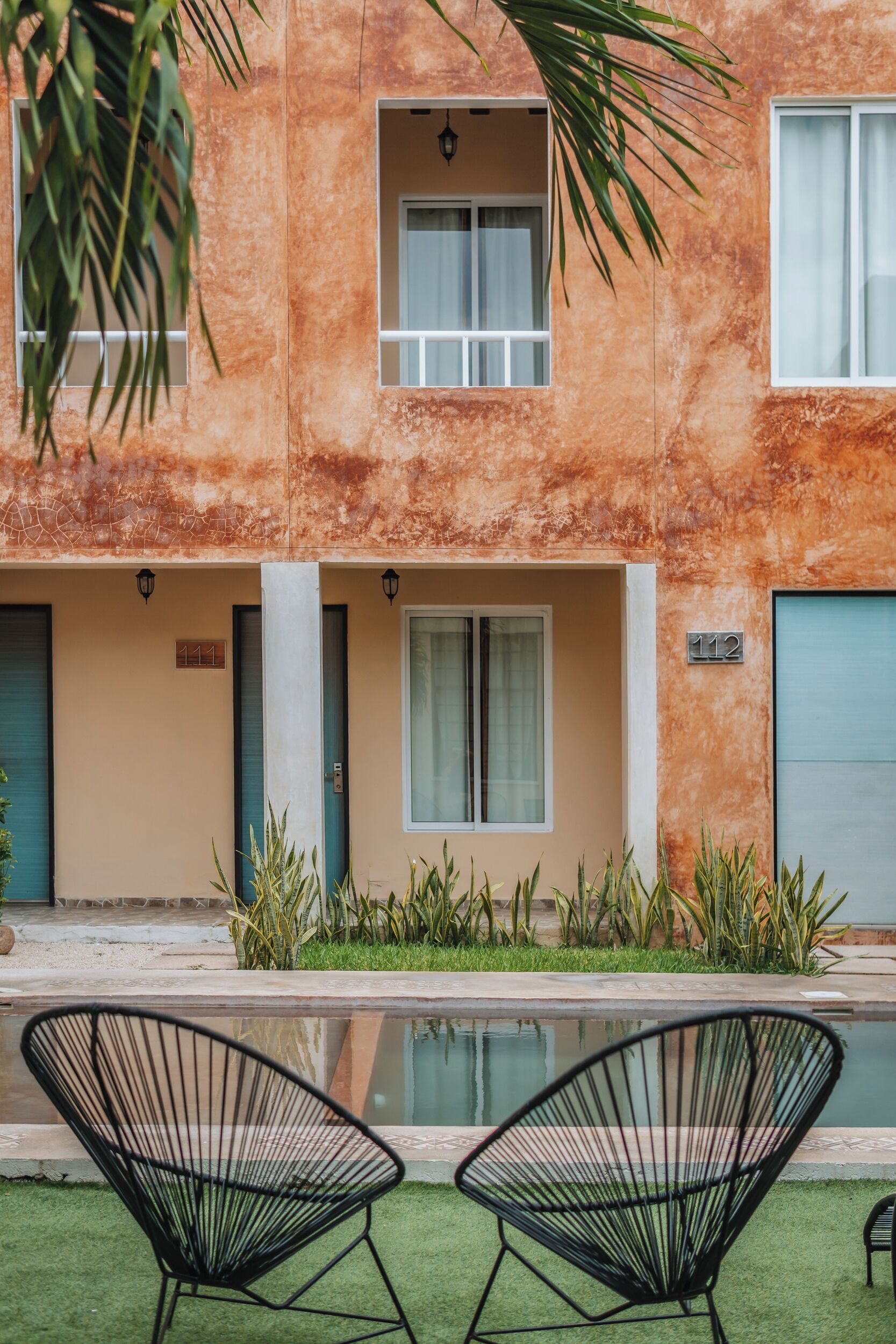 A modern building with a textured orange facade, numbered 111 and 112, featuring a small pool and two black wireframe chairs in the foreground.