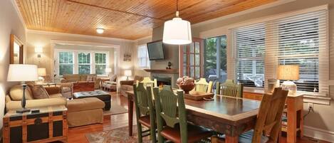 A spacious living room with a wooden ceiling, a dining area with a wooden table and chairs, and a cozy seating area with a sofa and ottoman.