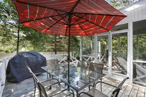 Une table à plateau en verre, des chaises en métal, un parasol rouge et un barbecue sur une terrasse en bois.