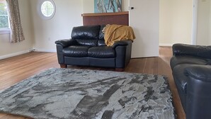 Un salon avec un canapé en cuir noir, un tapis à longues mèches gris, un tableau représentant des mouettes et une porte.