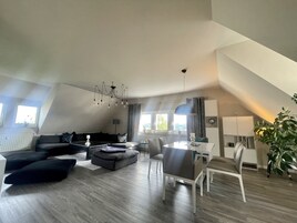 Amplia sala de estar con un juego de sofás, mesa de comedor y sillas, una araña y un ventanal con cortinas.