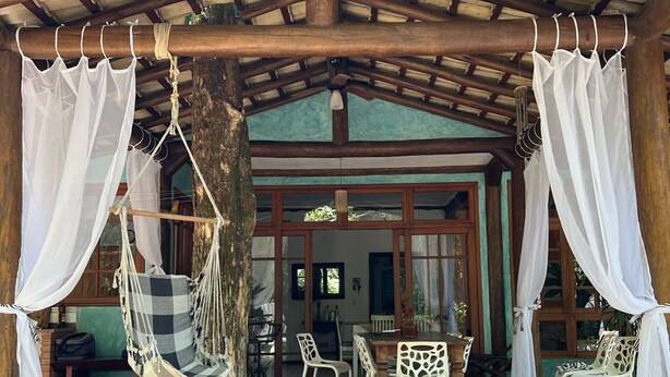 Un espacio al aire libre con techo, con piso de madera, sillas blancas y una hamaca.