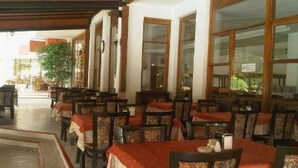 Een restaurant met meerdere tafels en stoelen, grote ramen en een plant op de achtergrond.