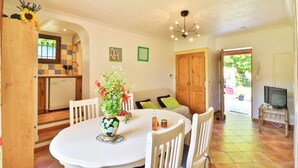 A dining area with a white table and chairs, a vase with flowers, and a kitchen in the background. - RENTALS-06 NOELIE'S HOUSE - 300M RIVIERE 30mn NICE CANNES ANTIBES (Le Bar-sur-Loup)