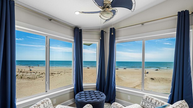 A room with a large bay window overlooking a beach, a ceiling fan, and patterned furniture.
