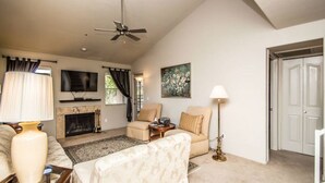 A living room with a fireplace, a ceiling fan, a television, and a door. - Great North Scottsdale Condo! (Scottsdale)