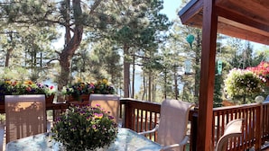 An outdoor patio with a glass-top table surrounded by wicker chairs, a wooden deck, and potted plants with flowers. - Views | Hot Tub | Fireplace | Scenic Serenity (Angel Fire)
