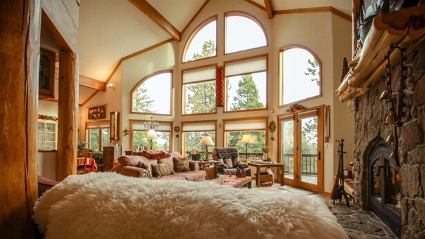 A spacious room with a large bed, a fireplace, and a dining area with a view of trees through large windows. - Views | Hot Tub | Fireplace | Scenic Serenity (Angel Fire)