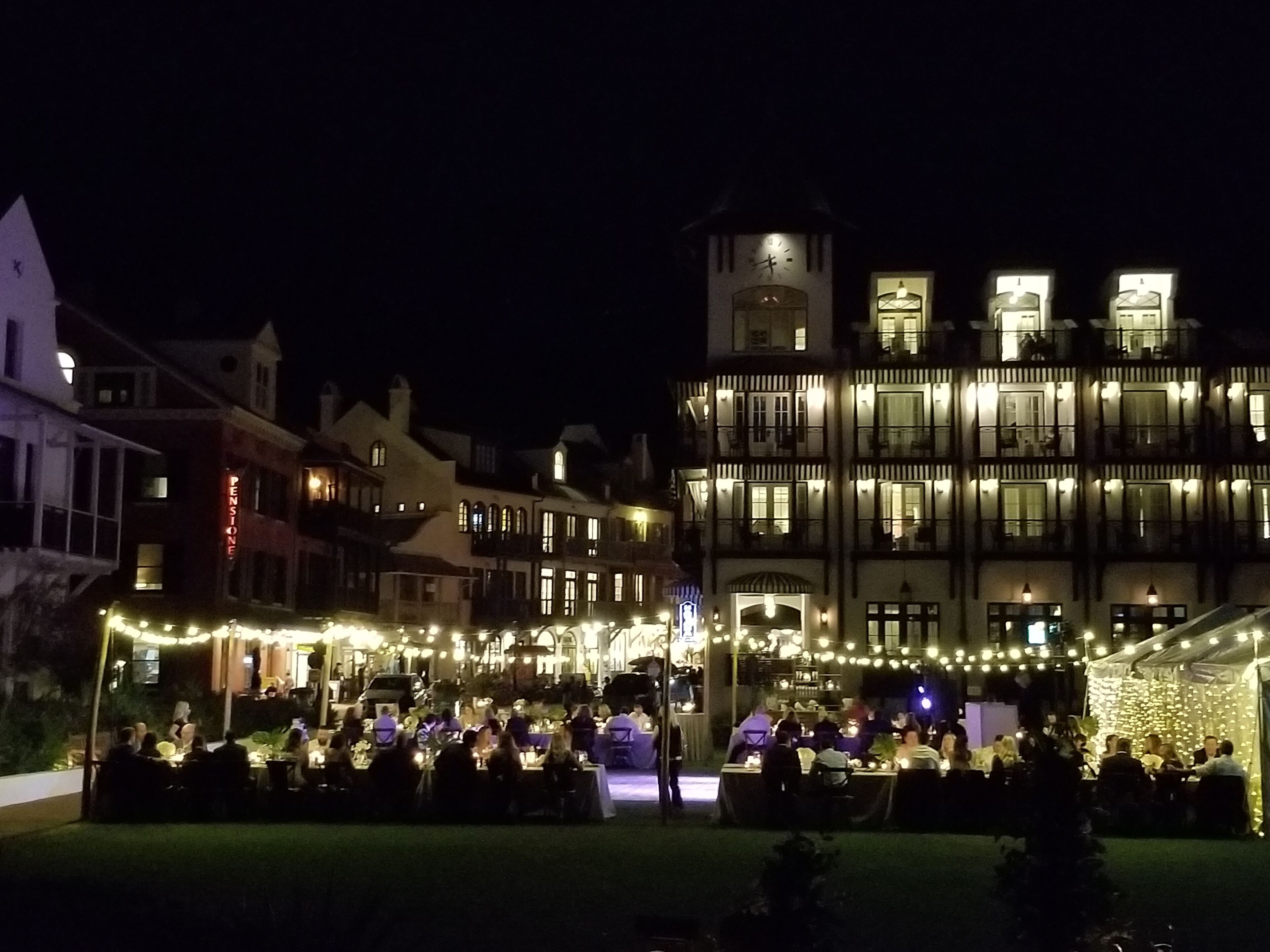 A historic building with multiple balconies and a clock tower, surrounded by string lights and outdoor dining areas.
