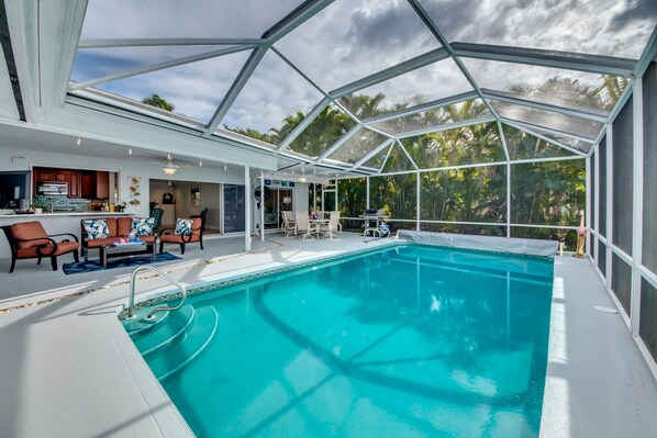 Una piscina interior con techo de vidrio, rodeada de sillones de descanso y una zona de cocina.