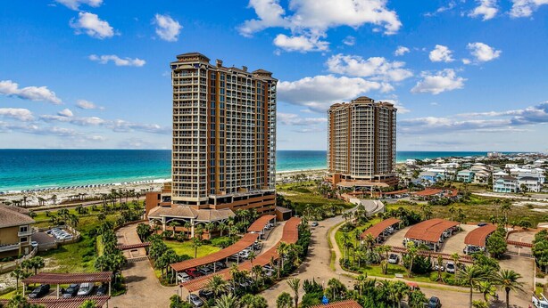 Een resort met twee hoge gebouwen, een strand en een parkeerplaats met palmbomen.