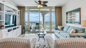 Een ruime woonkamer met een grote televisie, comfortabele zitplaatsen, een glazen salontafel en een balkon met uitzicht op de oceaan.