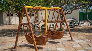 Un columpio de madera con dos asientos, un patio con baldosas y sillas verdes al fondo.