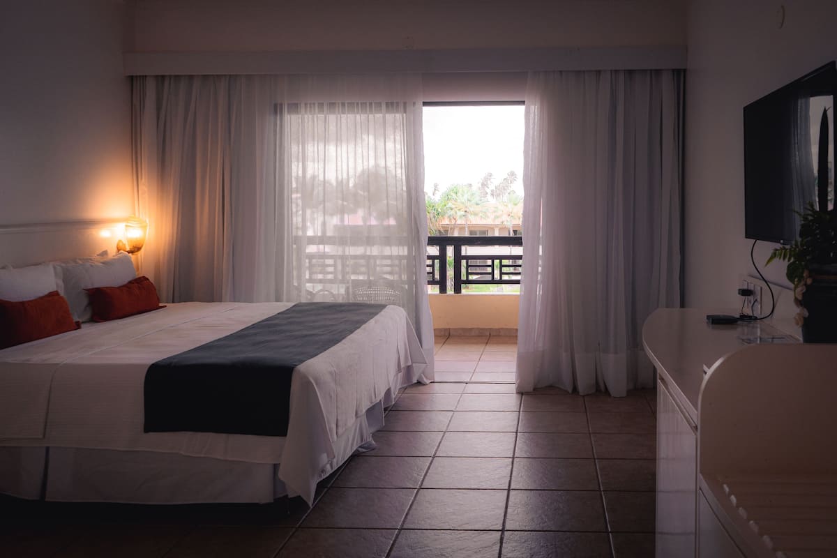 A hotel room with a bed, a desk, a television, and a balcony with curtains.