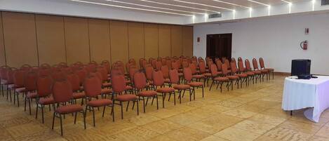 A conference room with rows of red chairs, a white table with a projector, and a tiled floor.