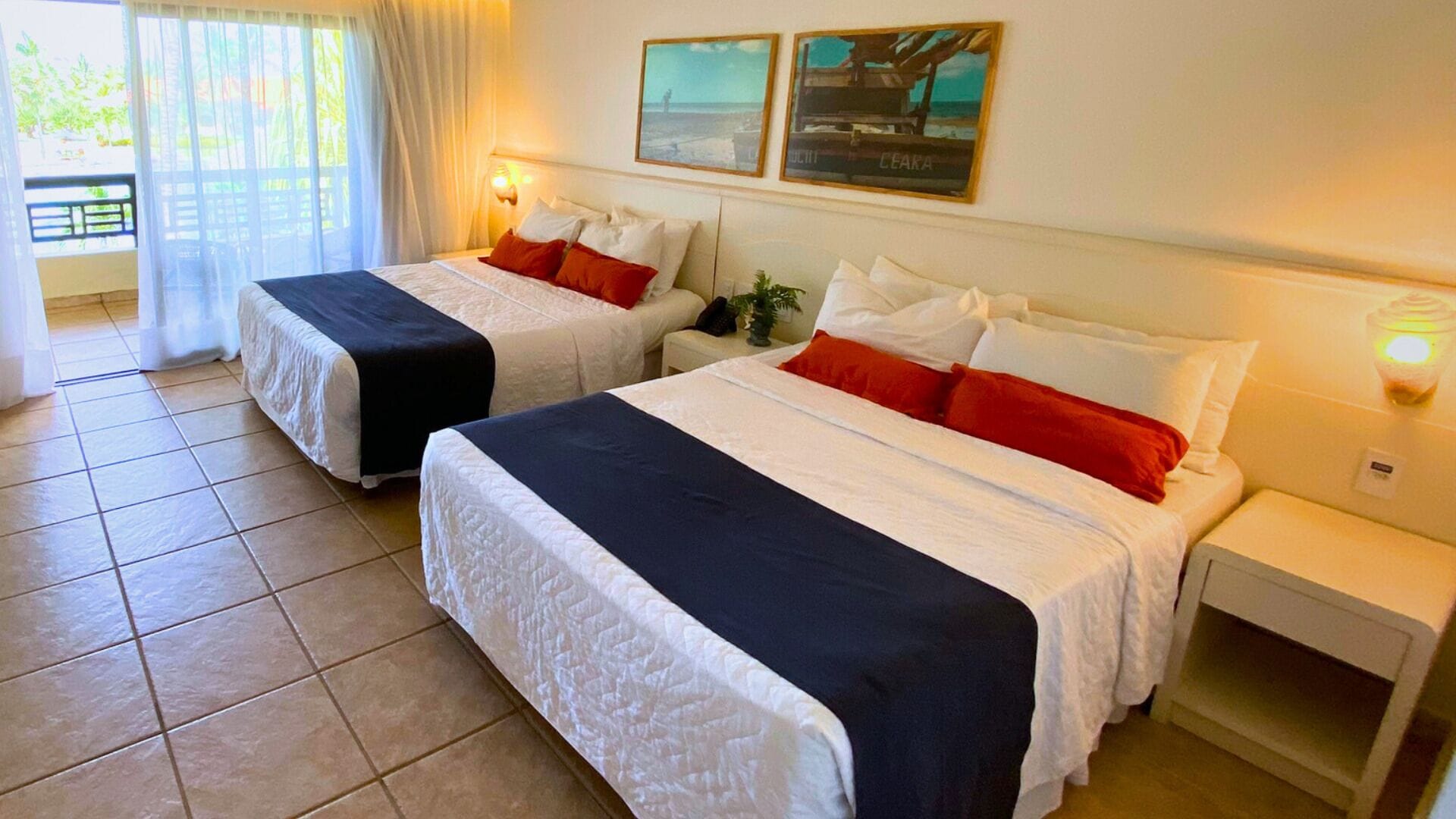 A hotel room with two beds, each with white bedding and red pillows. There are two bedside tables with lamps, and a window with curtains.