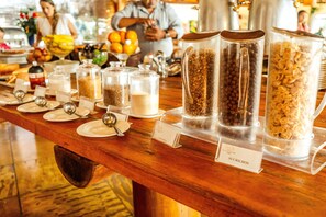 Une table de buffet proposant divers aliments et contenants étiquetés avec des noms tels que « Granola » et « Sucrilos ».