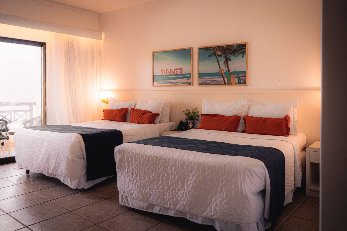 A hotel room with two beds, white bedding, orange pillows, a view of the sea, and a painting of a beach scene.