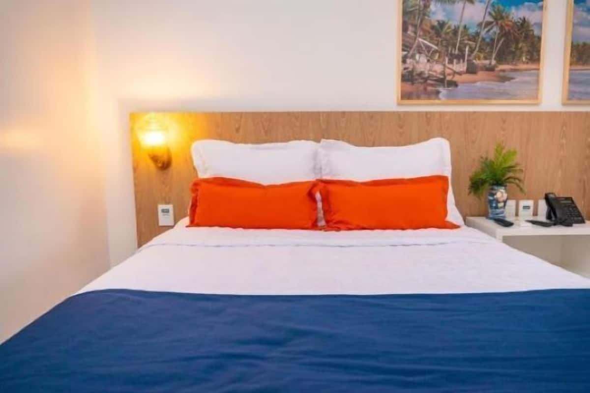 A neatly made bed with white and orange bedding, a wooden headboard, and a nightstand with a phone and a small plant.