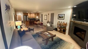 A modern living room with a sofa, coffee table, and fireplace. There is a kitchen area in the background with wooden cabinets and a dining table. - August Special, Great Rates! Locking bike storage! (Gunnison)