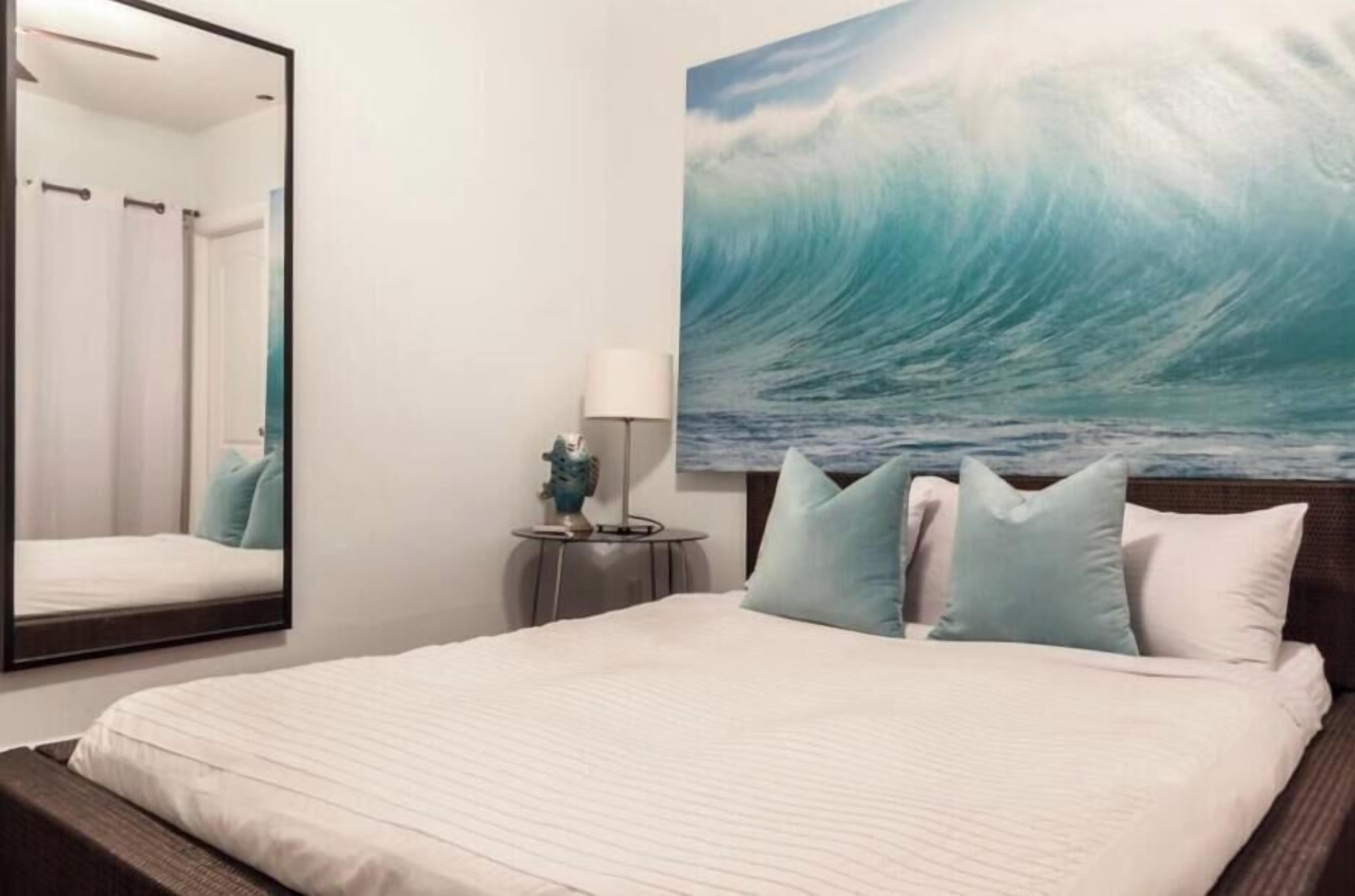 A bedroom with a large bed, a nightstand, a mirror, and a wall-mounted artwork of a wave.