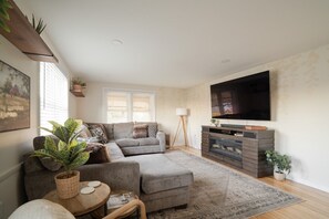 A living room with a grey sofa, a television, a fireplace, and a large window with blinds.