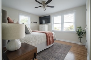 A bedroom with a bed, bedside table, lamp, TV, dresser, windows, and a ceiling fan.