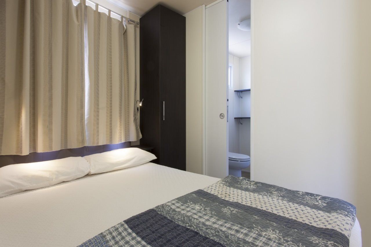 Une chambre à coucher avec un lit, une armoire sombre et une salle de bain visible à travers une porte ouverte.