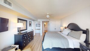 Une chambre à coucher comprenant un lit, des tables de chevet, une commode et un ventilateur de plafond, et une salle de bain visible à travers une porte ouverte.