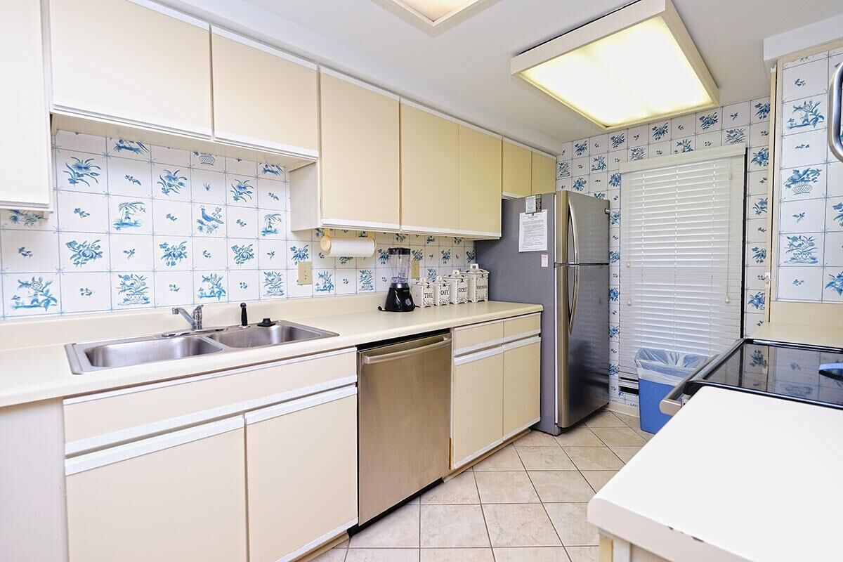 Una cocina con armarios color beige, un lavamanos de acero inoxidable, lavavajillas, refrigerador y una ventana con persianas.