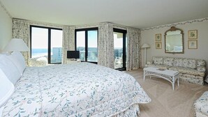 A hotel room with a large bed, a sofa, a small table, and a TV. The room has large windows with curtains and a view of the city. - Coastal Oasis Inviting 3BR Condo with Resort Pool (Pawleys Island)