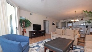 Ein Wohnzimmer mit blauem Sofa, beigem Sofa und einem Couchtisch. Ein Fernseher steht auf einem Holzgestell, und im Hintergrund ist eine Küchenzeile erkennbar.